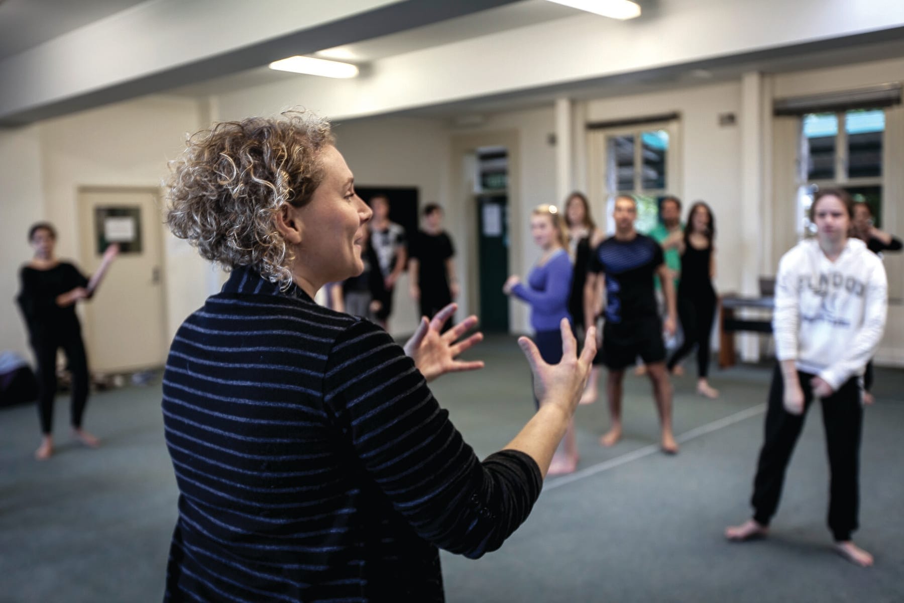 A tutor gesturing while leading a group of acting students standing in a rehearsal room during a voice class